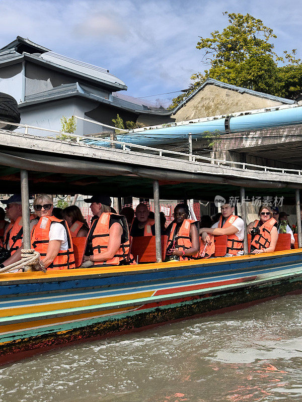 在Khlong Damnoen Saduak运河附近游览时，一艘带顶篷的木船载着穿着安全夹克的游客，这艘游船行驶在通往Tha Chin和Mae Klong河的水路上